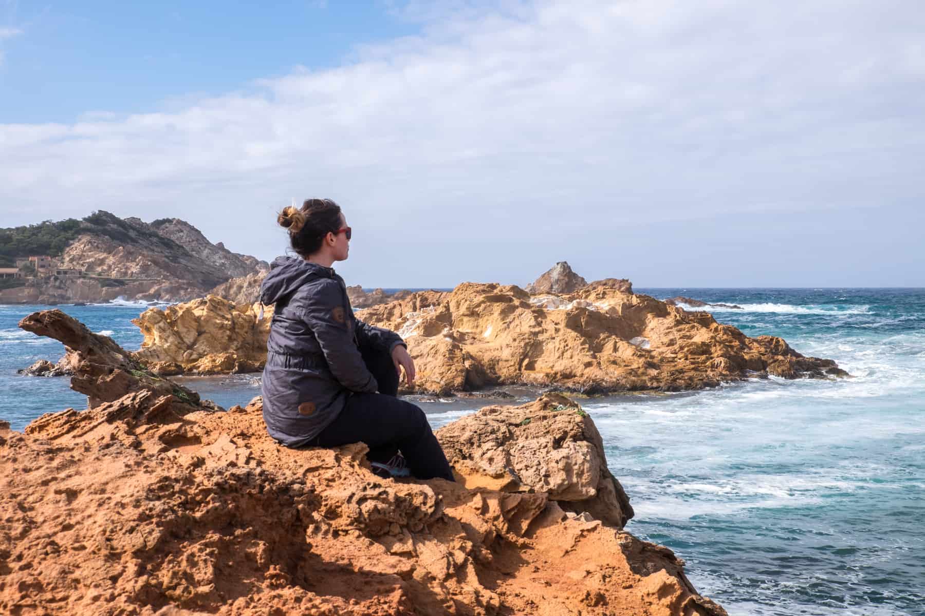 A woman in a navy blue coat sits on a jagged golden rock on the coastline of Menorca, Spain looking out to the ocean.