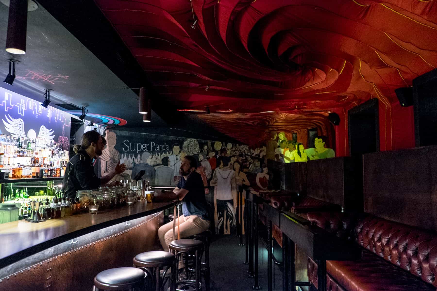 A man sits at a bar talking to two barmen. The bar is circus themed with painted artwork walls and a red, velvet ceiling. 