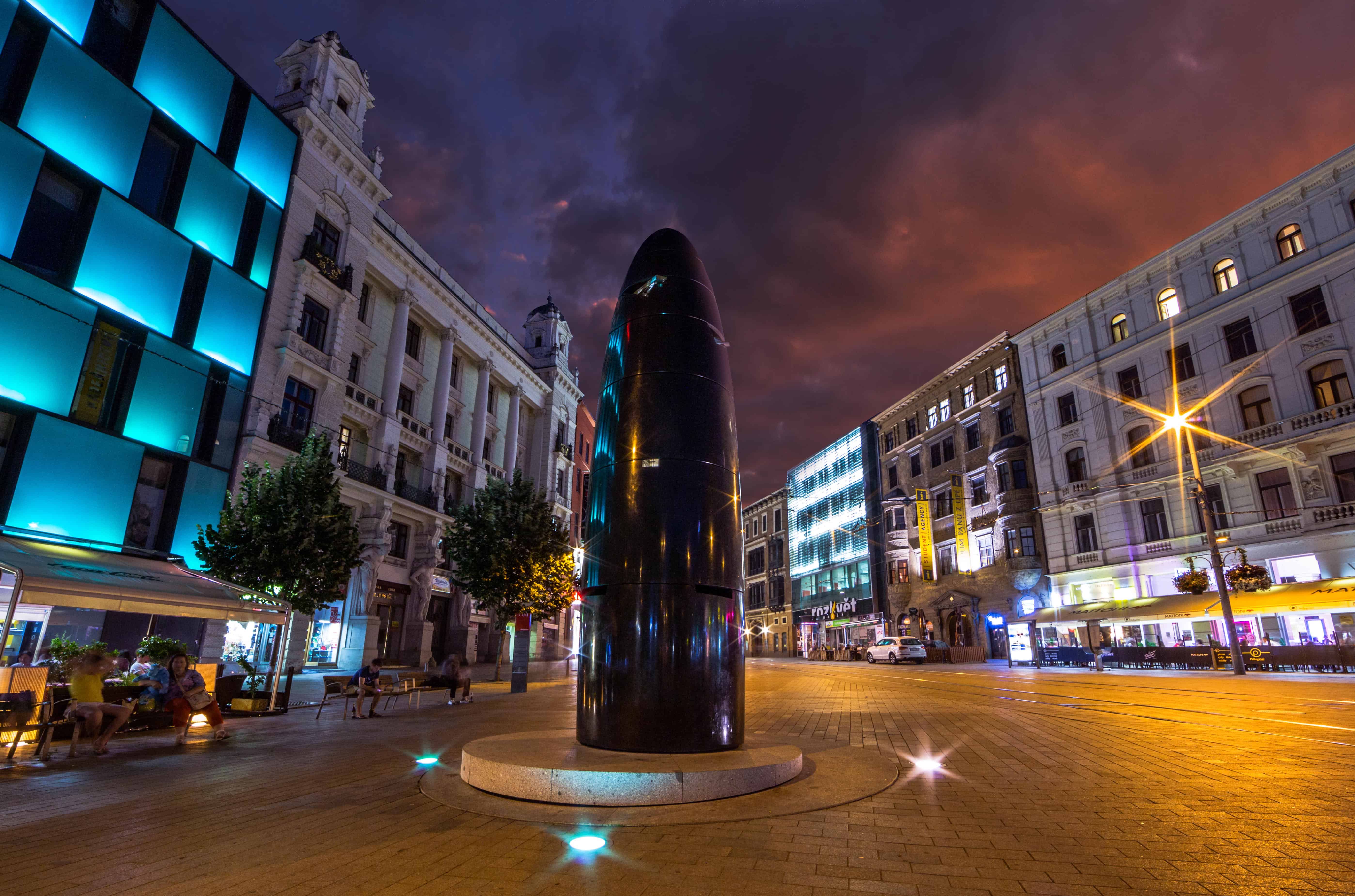 A large black phallic looking clock in Brno, Czech Republic. It stands in the middle of an avenue of tall buildings in various architectural styles. 