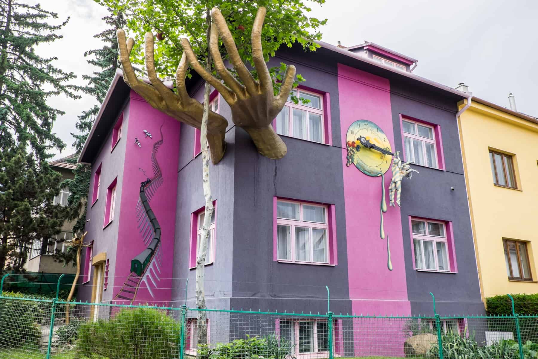 A purple and pink stripped building with a giant pair of golden hands protruding from it. A tree grows in between the hands and a figure can be seen hanging from a clock on the front wall. 