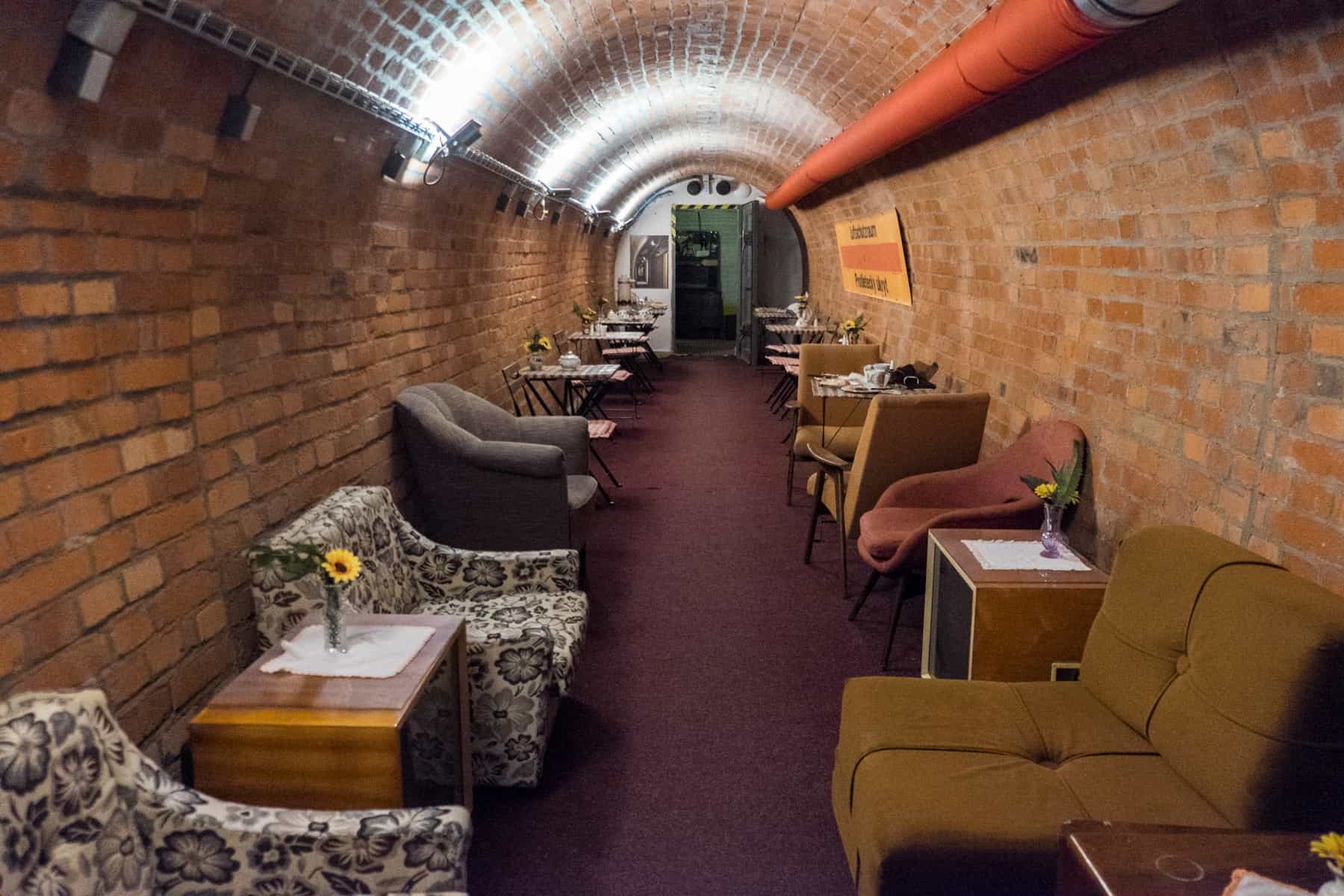 A long brick tunnel filled with an orange wall pipe, filled with old chairs and tables in muted colours - a lounge room at the bunker in Brno. 