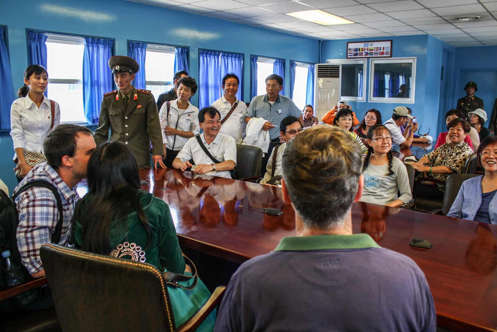 Tourists sit around a table in the Military Armistice Commission Conference Room when visiting the DMZ in North Korea. A North Korean guard in military green uniform is present. 