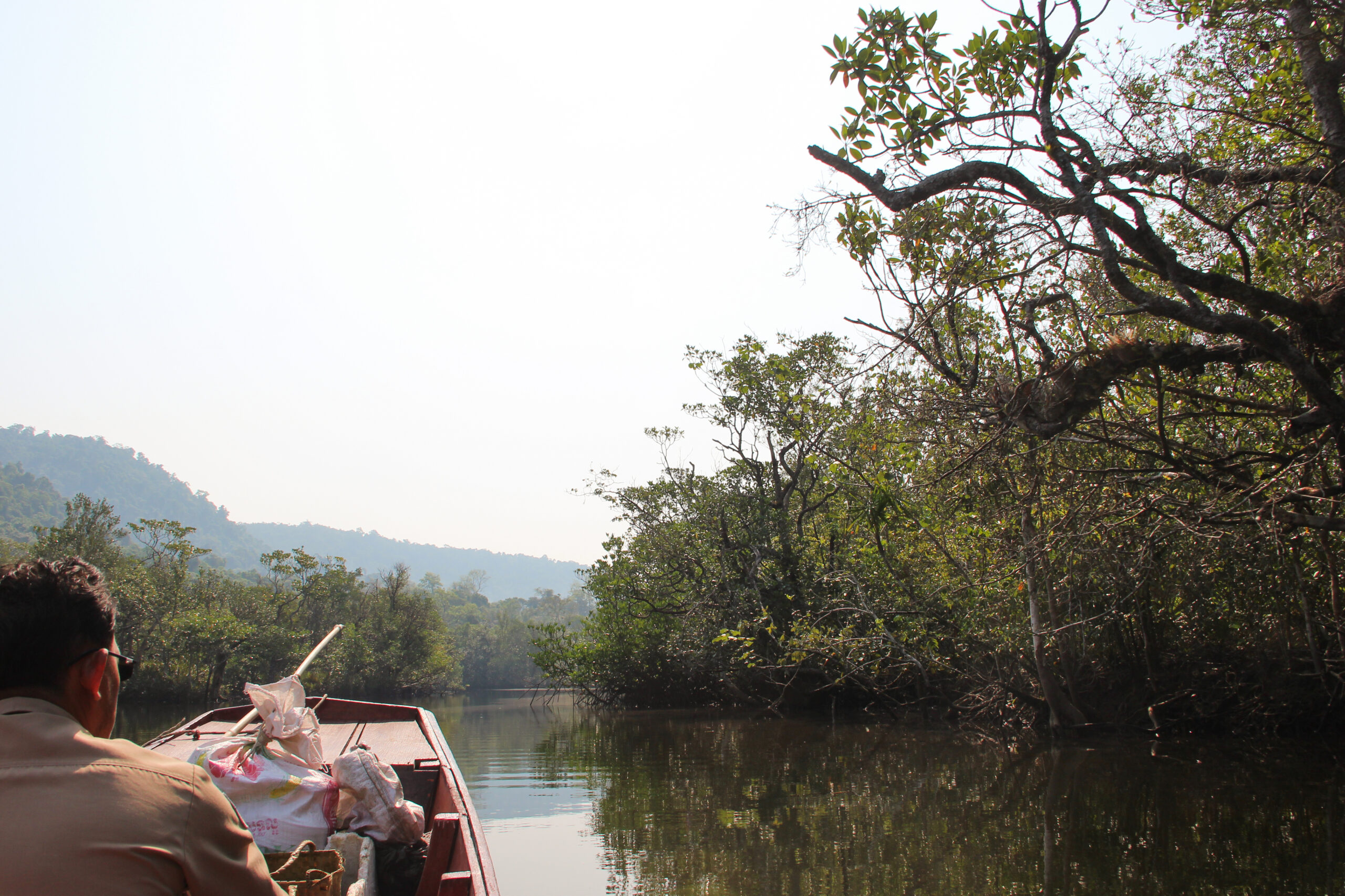 A man in a beige short sits at the front of a wooden boat, loaded with white bags, as it drifts through a forested river stretch backed by hills.
