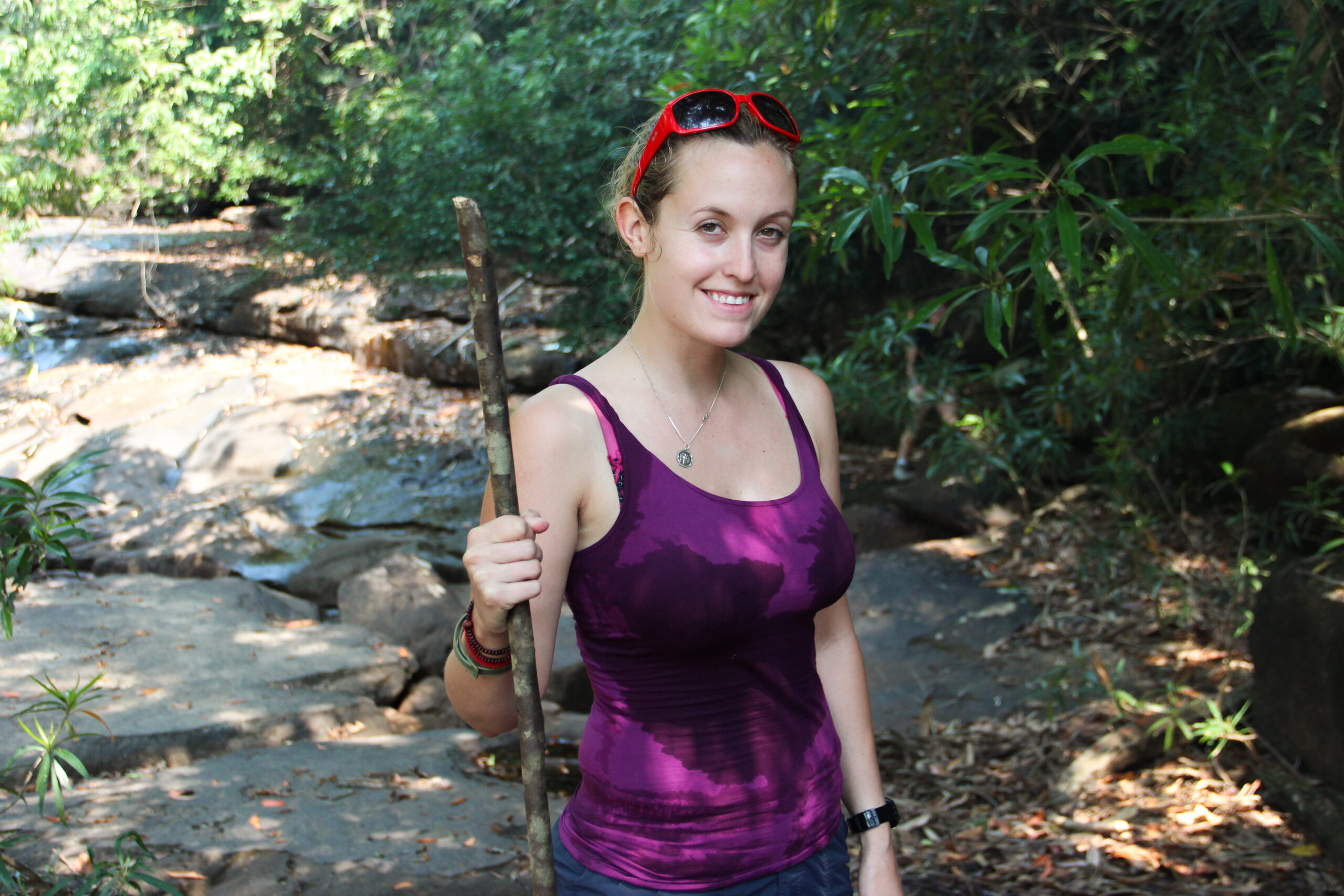 A woman in a water-soaked purple vest, red sunglasses on her head and holding a wooden stick, looks at the camera while trekking a rocky pathway through a jungle in Koh Kong Island.