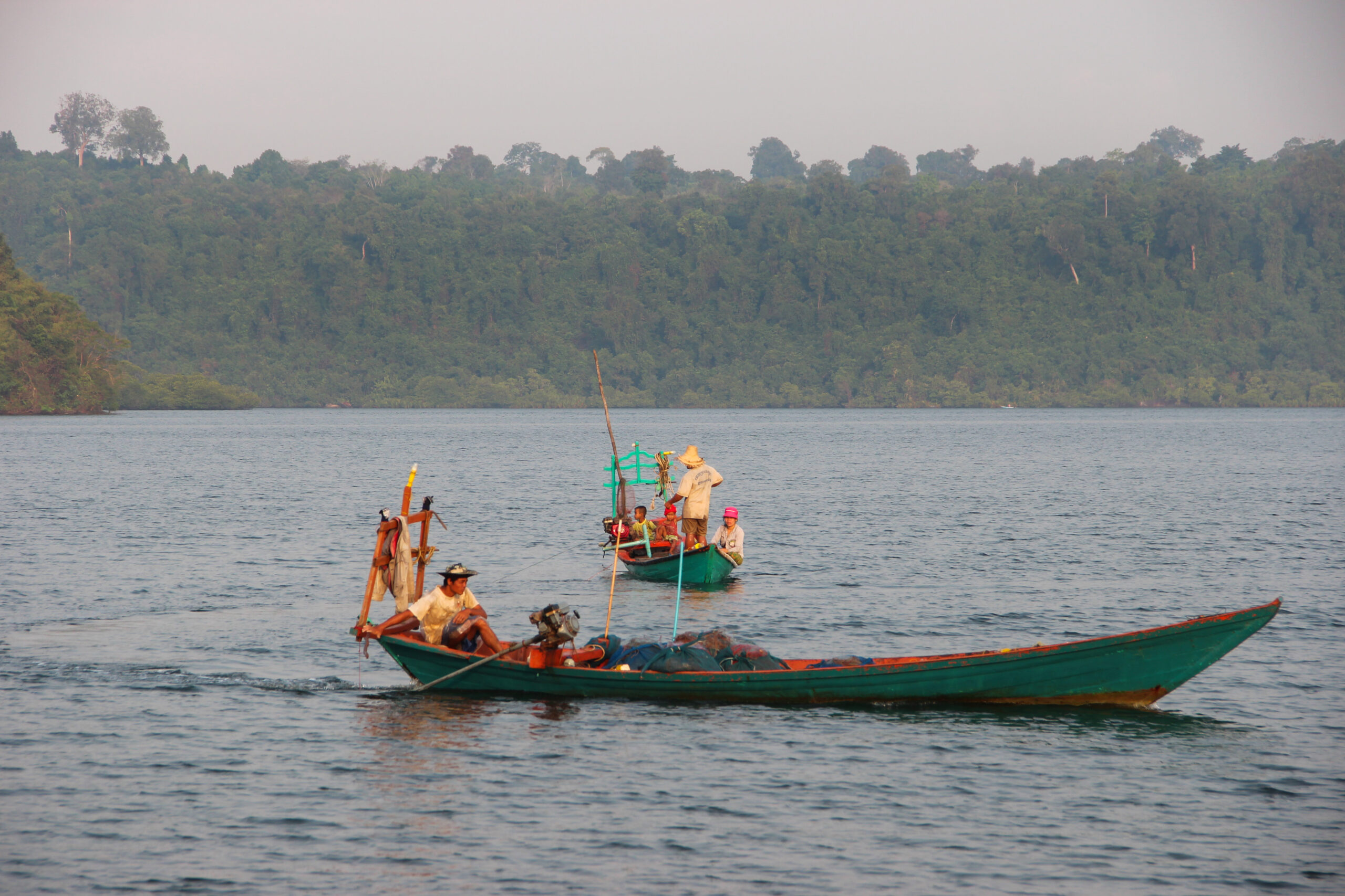 Two long and narrow teal hued fishing boats on the wide expanse of forest-backed water in Koh Kong province, Cambodia.