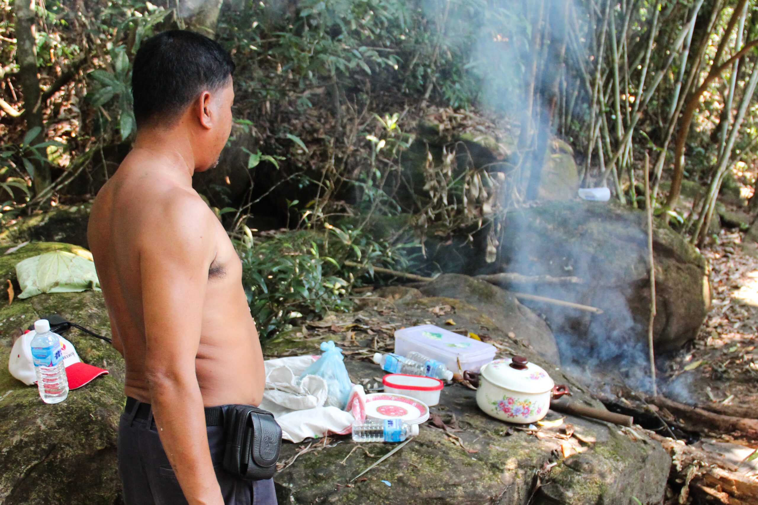 A topless man looking at a a makeshift kitchen and open fire in the jungle in Koh Kong island.