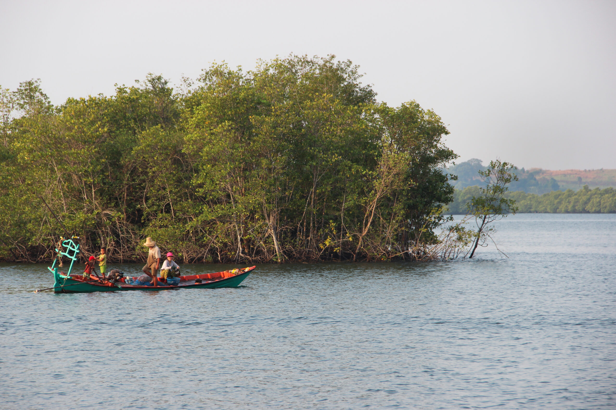 Two fisherman and a child on a small wooden boat on a wide body of water in front of a mangrove.