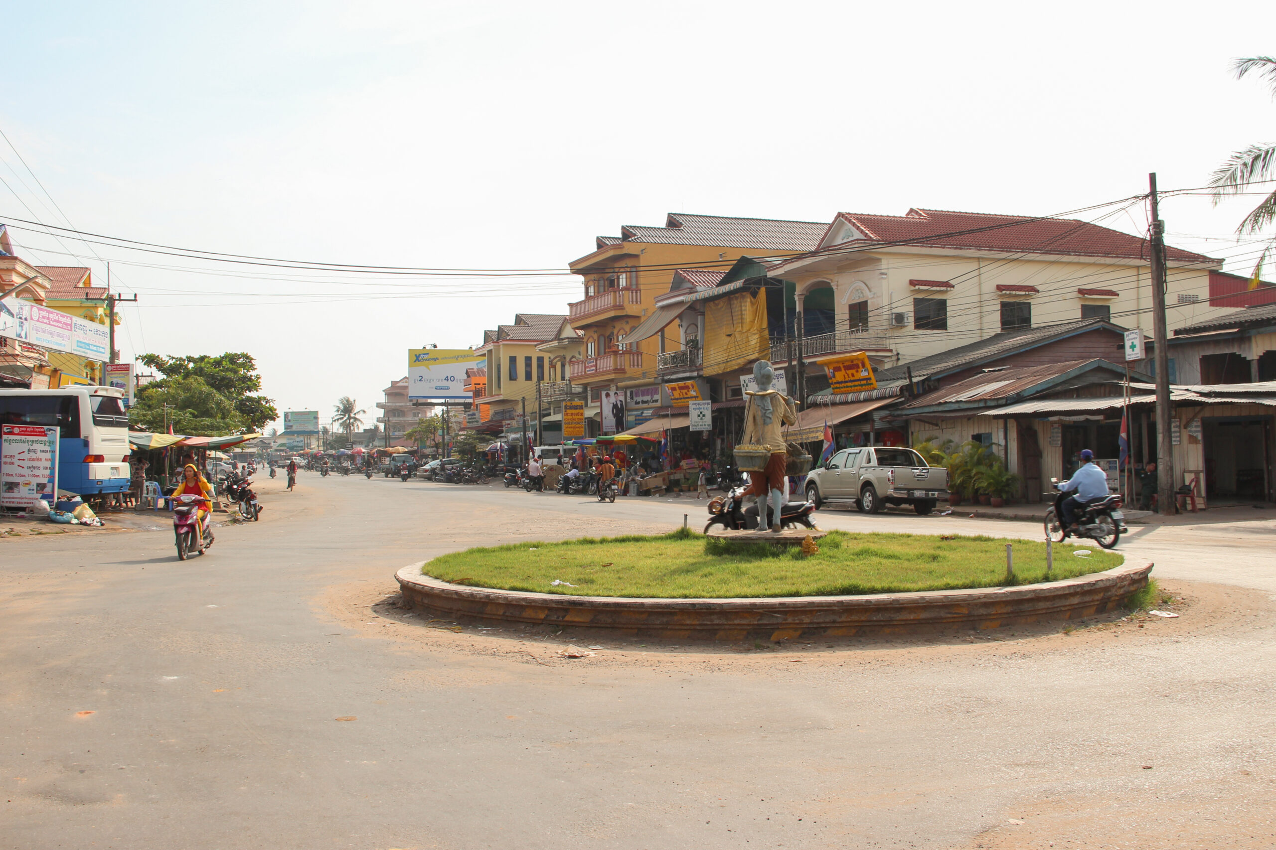 Motorbikes circle a tine grass-covered roundabout with a statue, in a small town with low-rise yellow buildings.