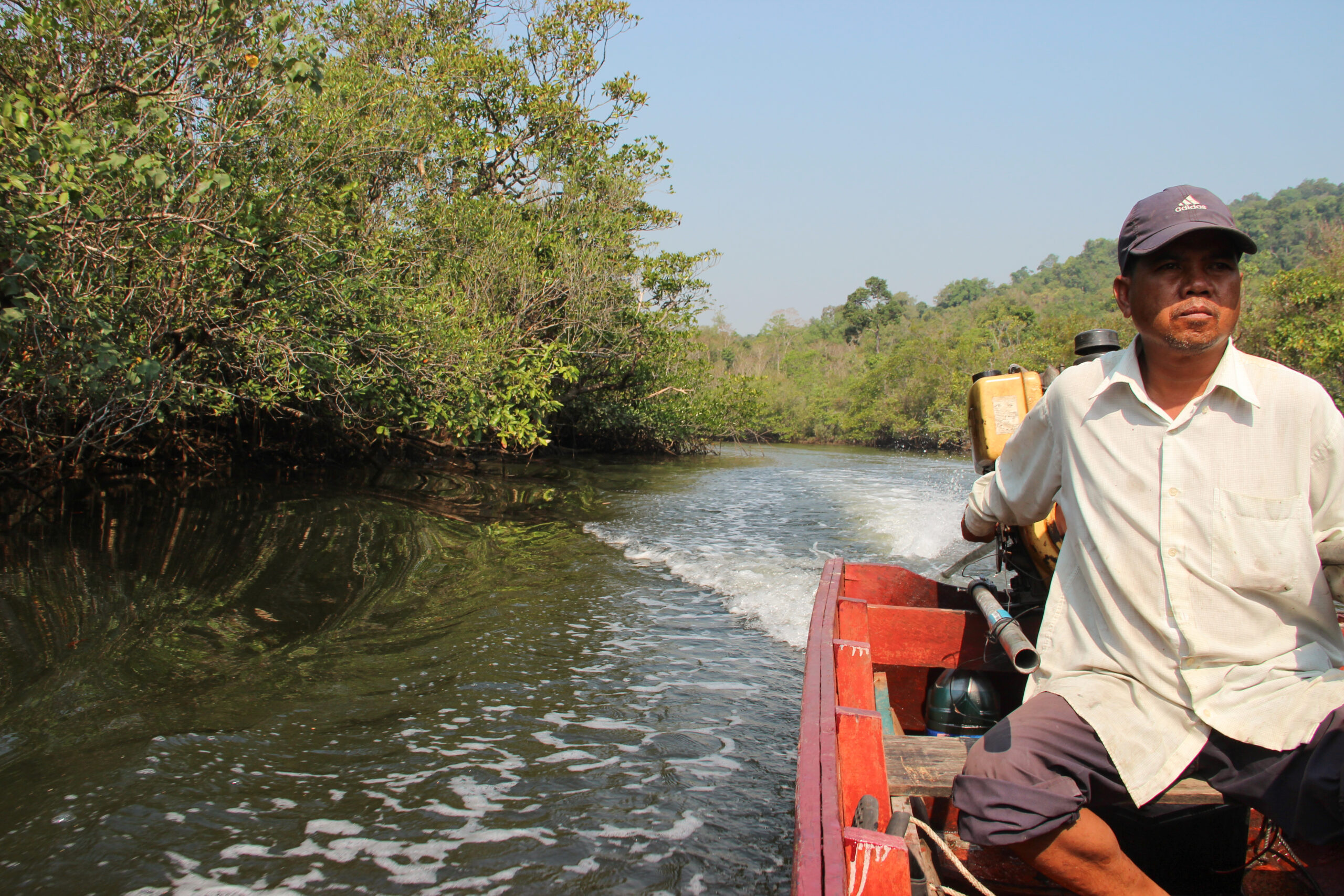 A man in a white shirt and dark cap driving an orange, wooden speed boat through a forested river in Koh Kong.