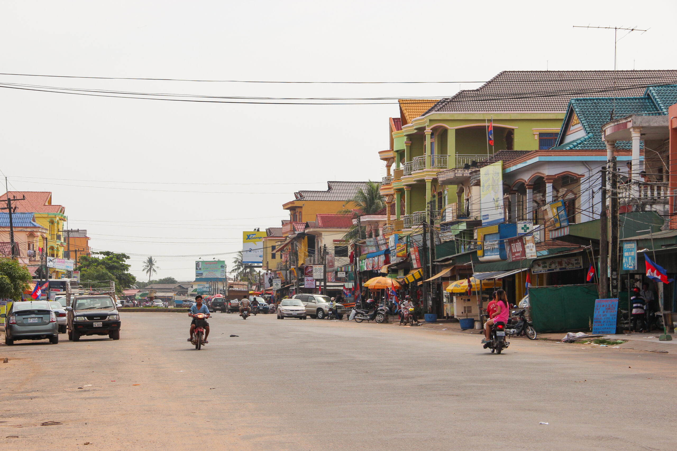 Motorcycles and cars on a wide, dusty road in Koh Kong town, lined with multicoloured low rise buildings.