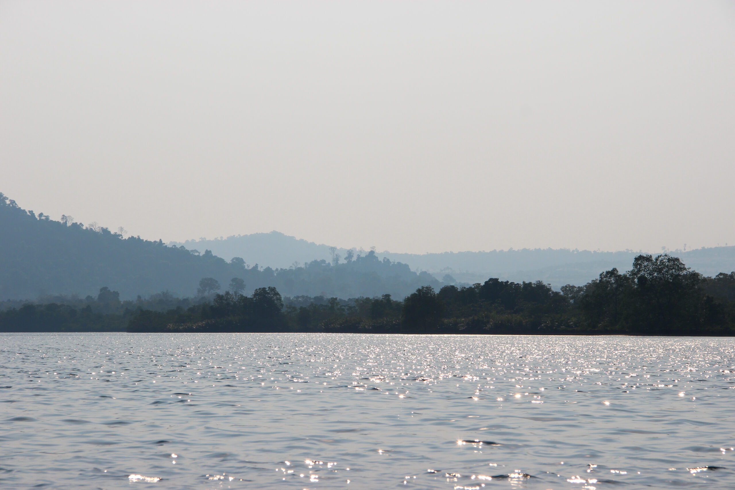 View from calm glistening waters towards a scenic three-layered forested and hilly landscape, in a dusky blue light.