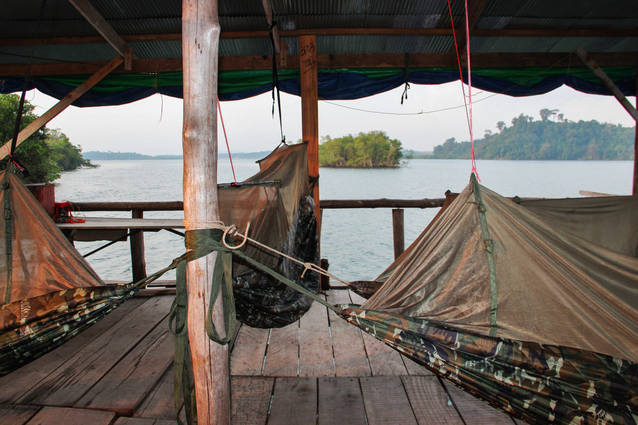 Four army camouflage hammocks on a covered wooden deck overlooking a body of water and green mangroves.