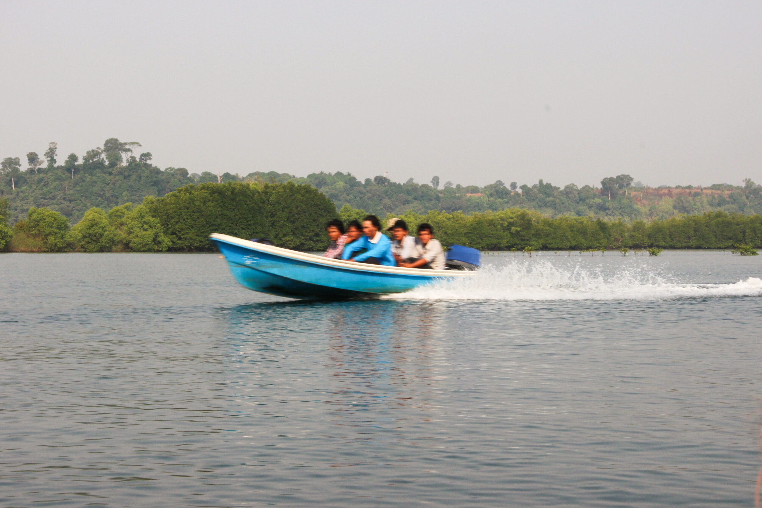 A small blue speed boat, whose six passengers are blurred in the picture by the speed, whips up some light white waves as it travels through a wide river backed by forest.