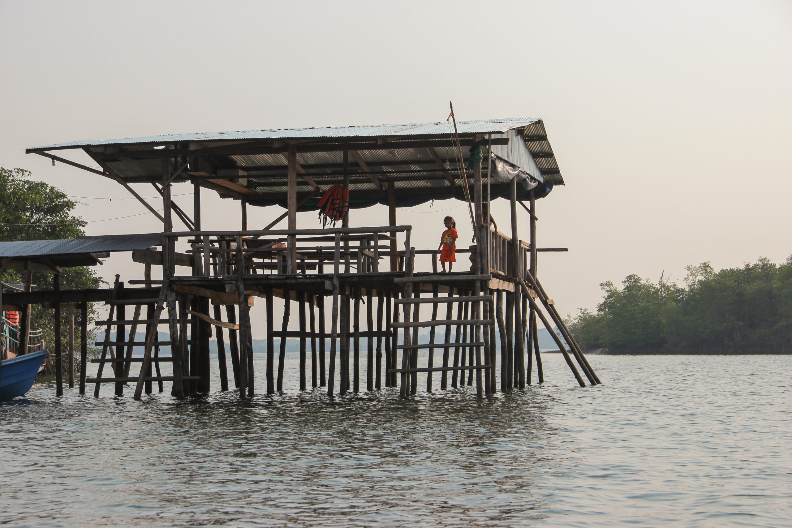 A child stands on the deck of a covered stilt house that stands in calm river waters surrounded by green groves.