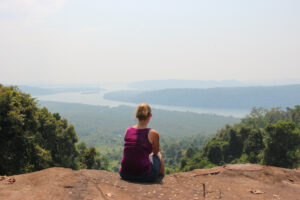 A woman in a purple vest sits on a sandy, ochre ledge overlooking a scenic view of a coastal shoreline on Koh Kong Island and the jungle-covered hills in the background.