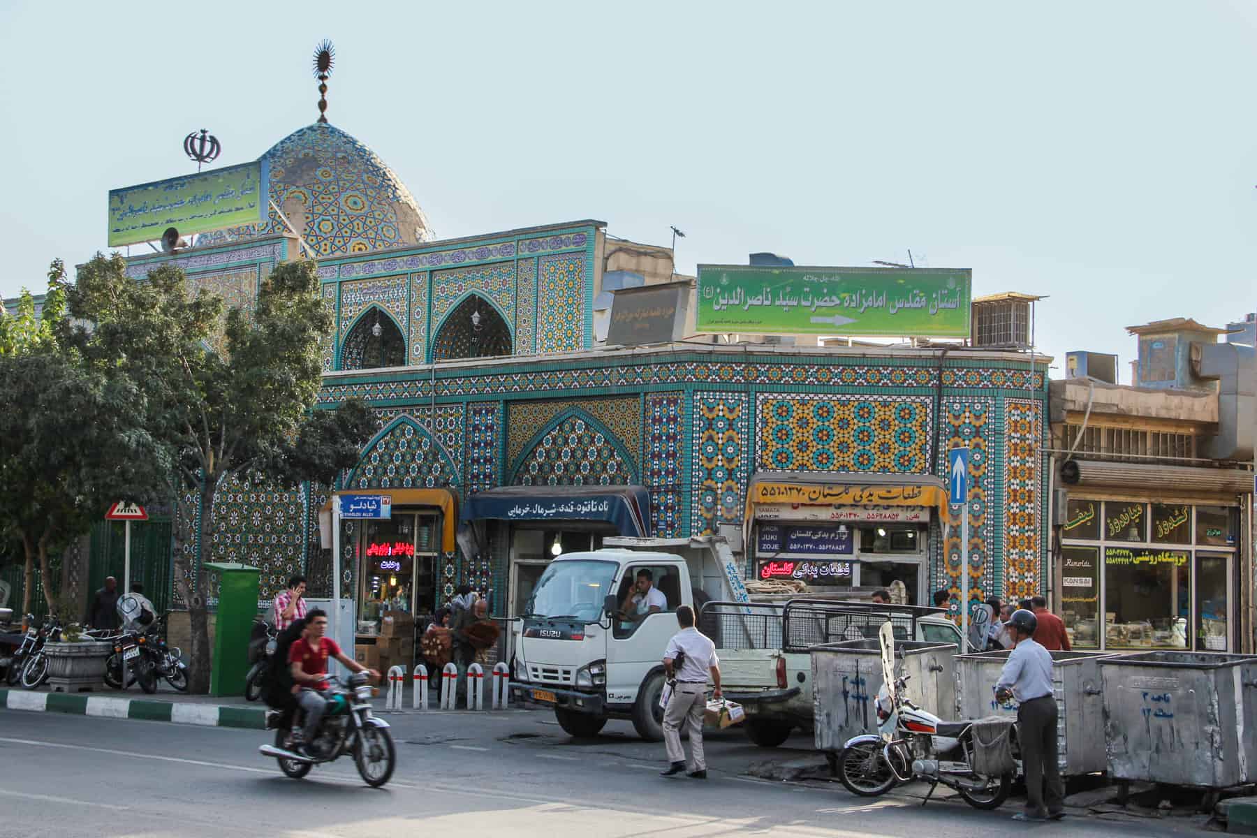A street scene in Iran showing people and traffic outside a blue and yellow mosaic covered building and dome. 