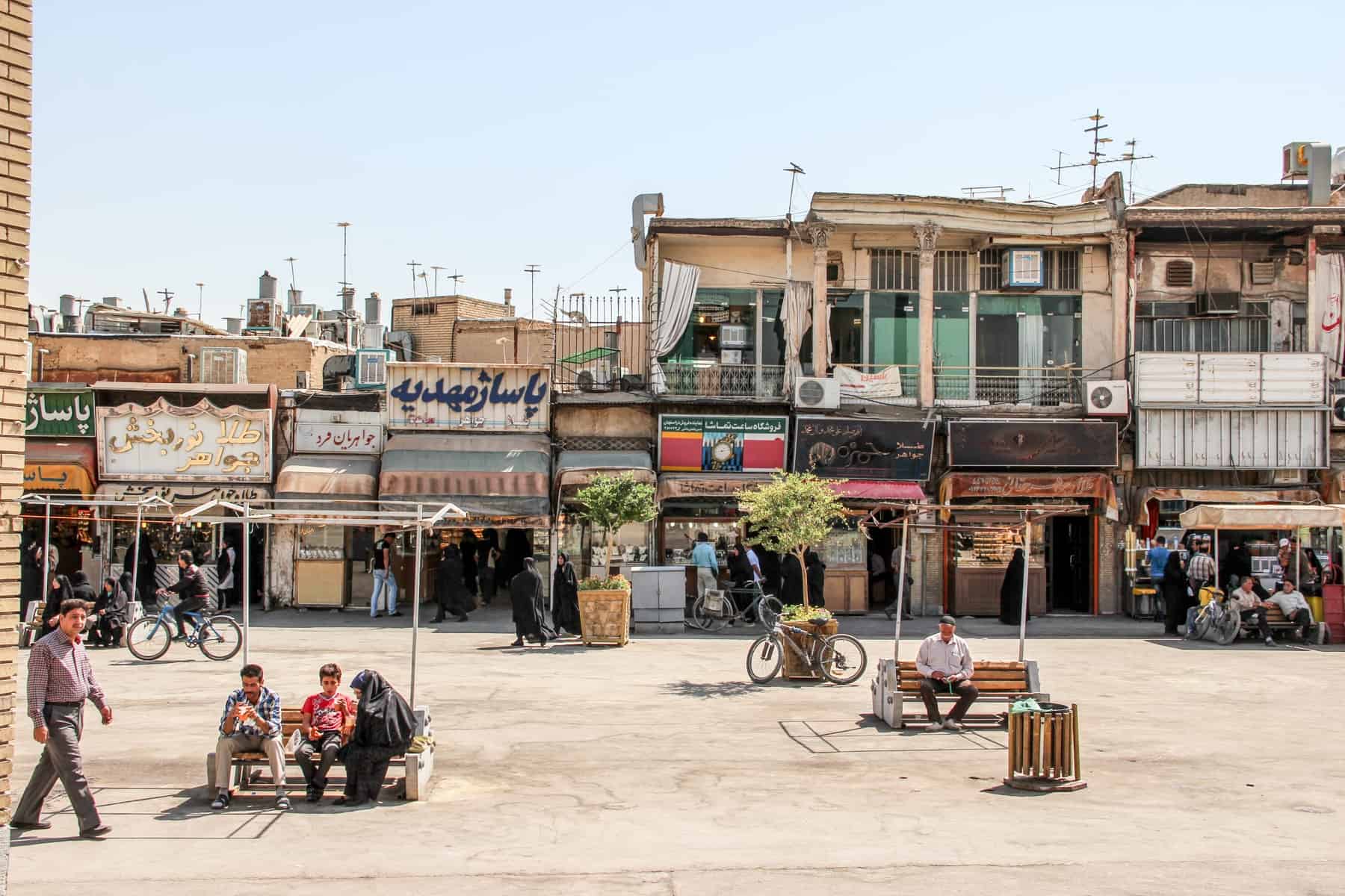 People sitting on benches on a beautiful street in Iran full of narrow, rectangular buildings and shop fronts. 