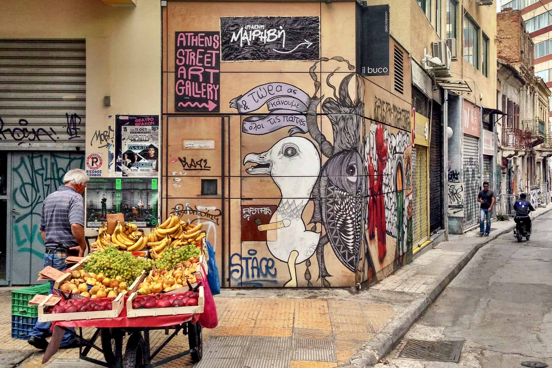 In Athens city, a man stands next to a cart full of bananas, grapes, pears and apples in front of a building covered in animal street art with a sign pointing to the 'Athens Street Art Gallery'. 