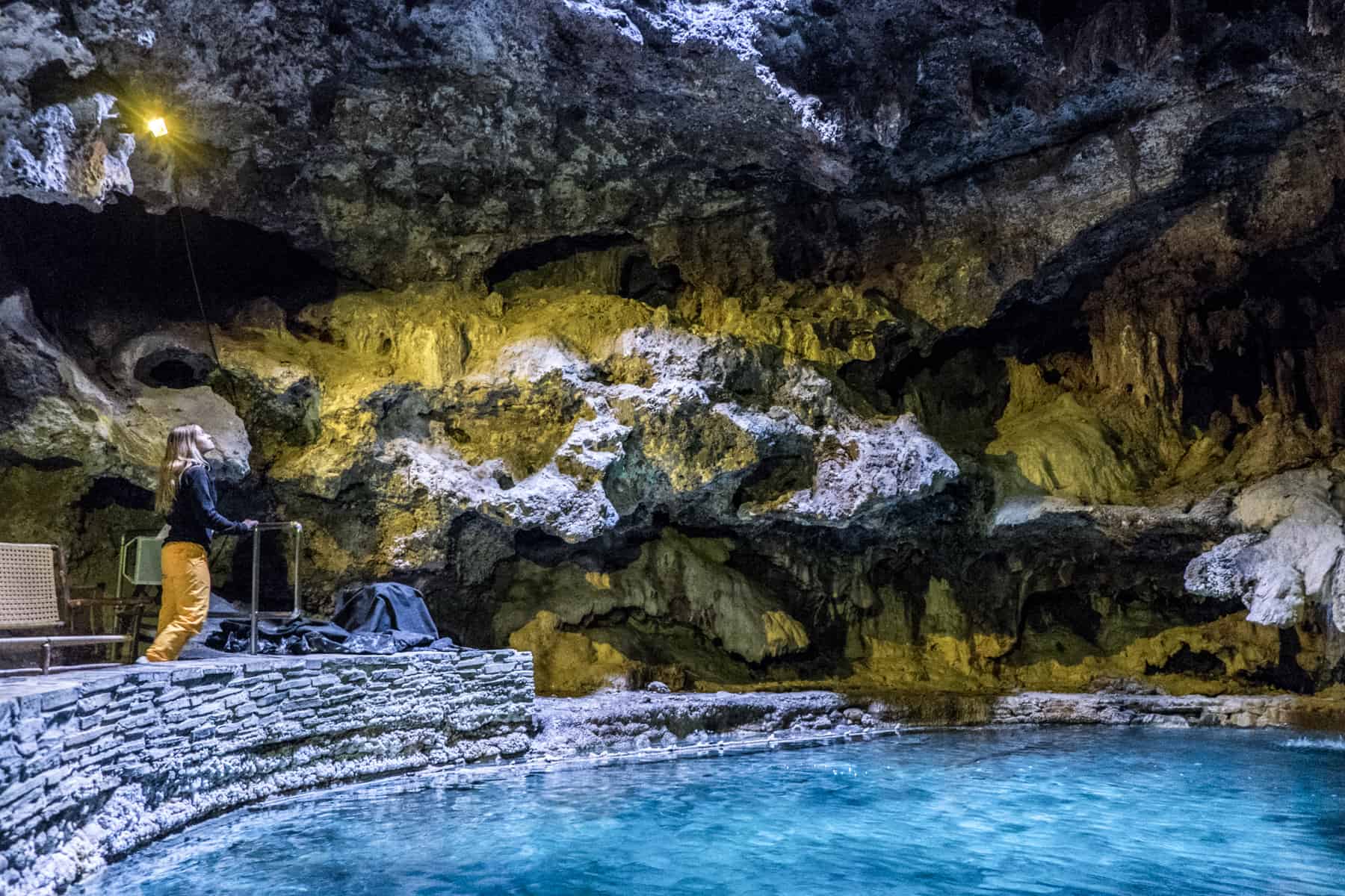 A woman in a black top and yellow trousers looks up towards the ceiling of a cave in Banff filled with a basin of bright blue water. 