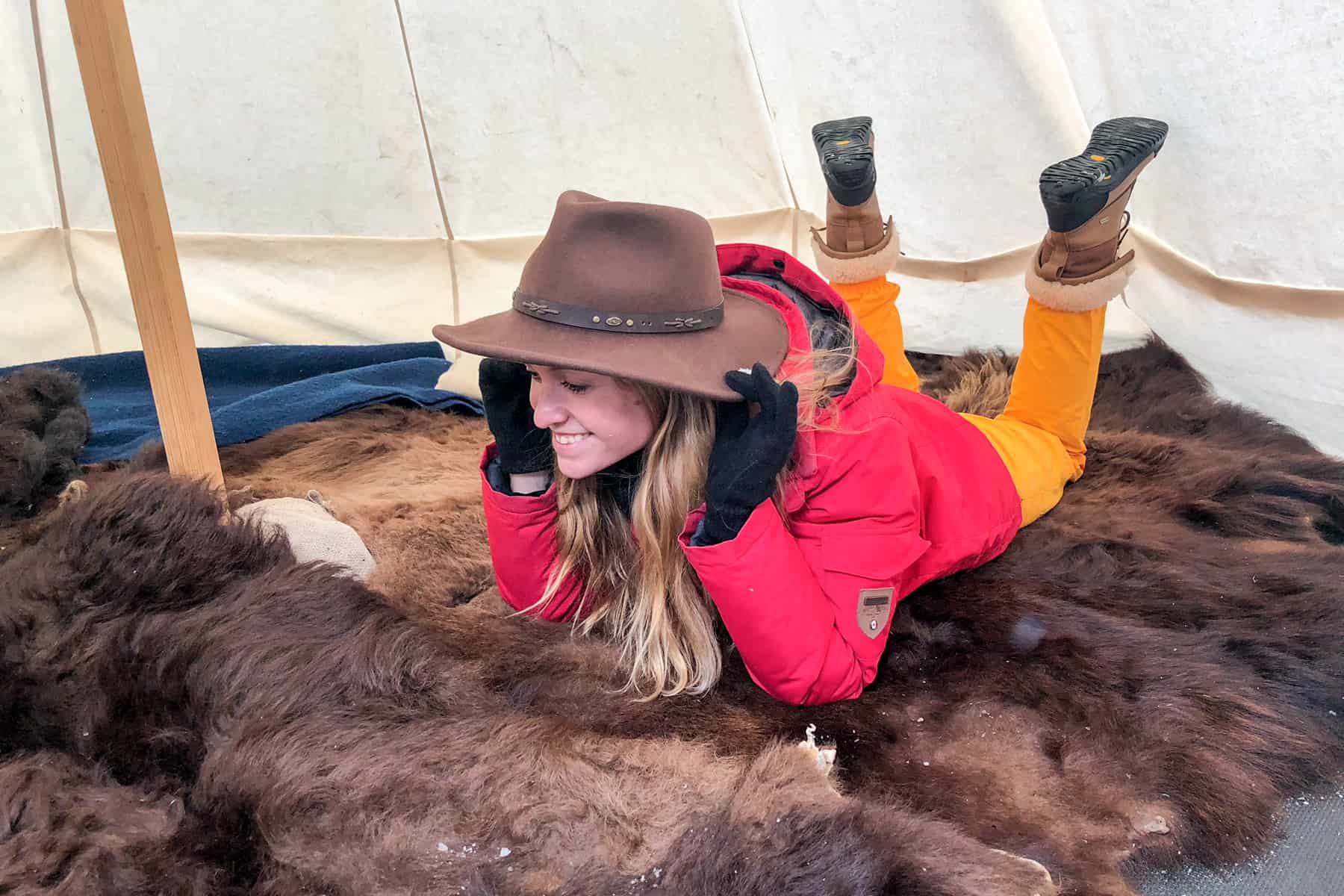 A woman in a red coat and yellow trousers wearing traditional Canadian hat while laying on rugs of fur. 