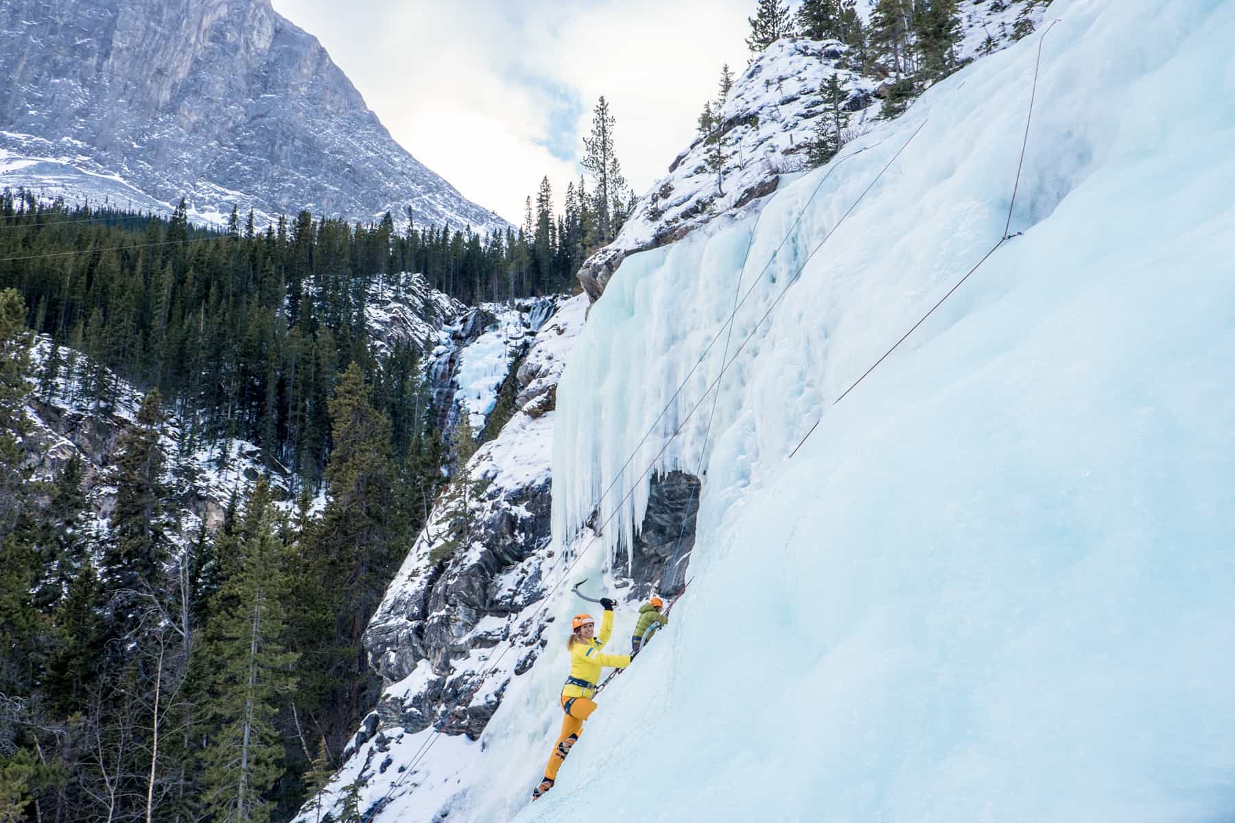A woman in a yellow jacket and orange pants ice climbing a frozen blue-ice waterfall in winter in Banff.