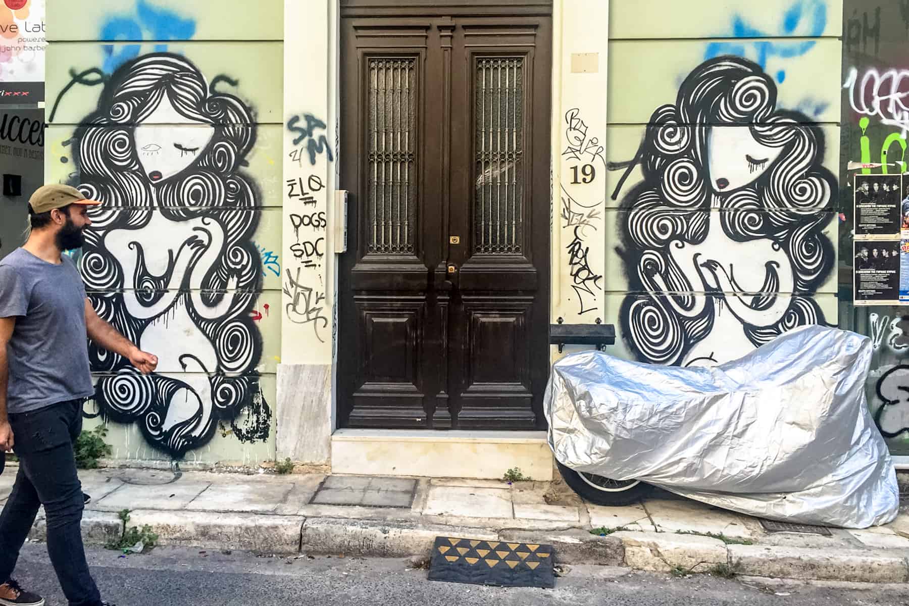 A man walks past a mint green building with a brown door that covered in black and white swirl style street art in Athens. 