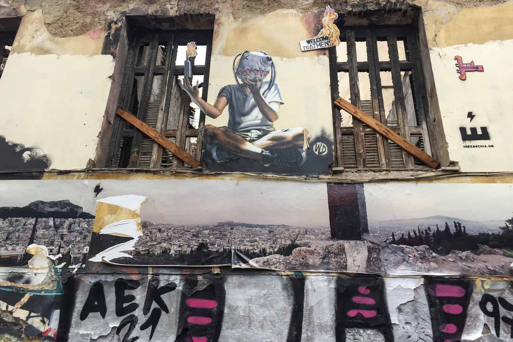 Athens street art on an abandoned building showing a guy in a mask, sitting crossed legged holding a molotov cocktail. Below is a panoramic photograph of the city. 