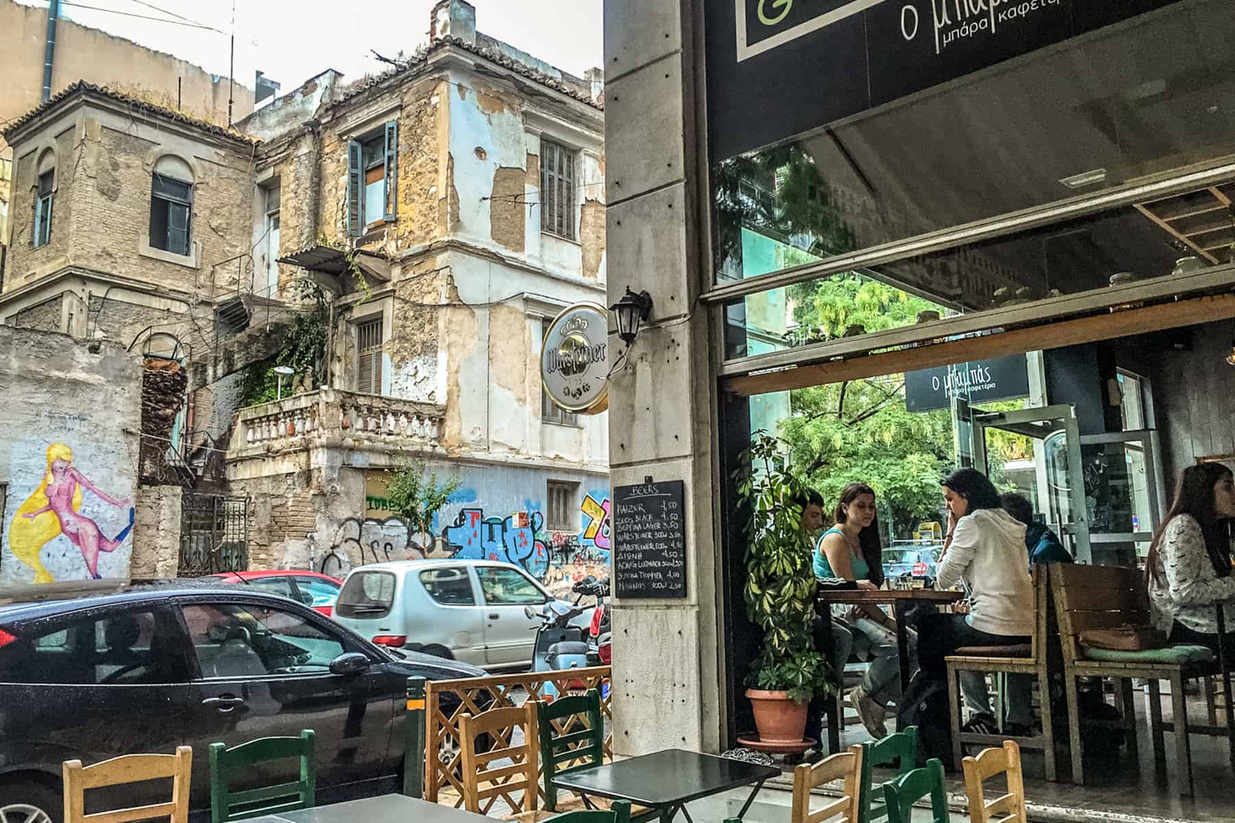 People sit in a cafe in Athens opposite a crumbling building covered in street art.