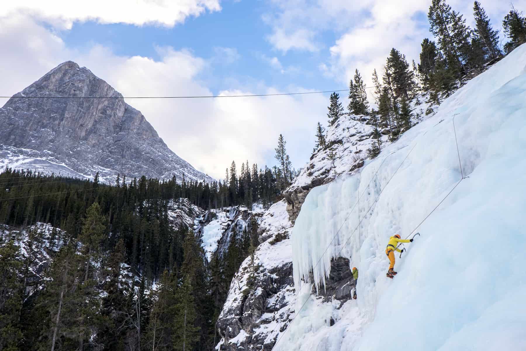 An ice climber in yellow and orange clothing scales a frozen waterfall to a backdrop of forest and mountains - one if the things to do in Banff in winter. 