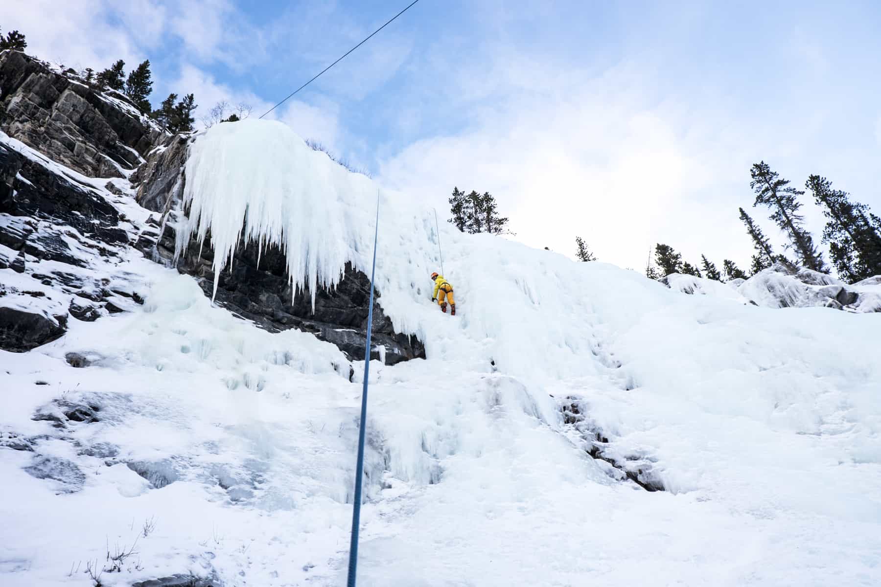 A woman in yellow and orange clothing climbing to the top of a frozen waterfall in Banff in winter.