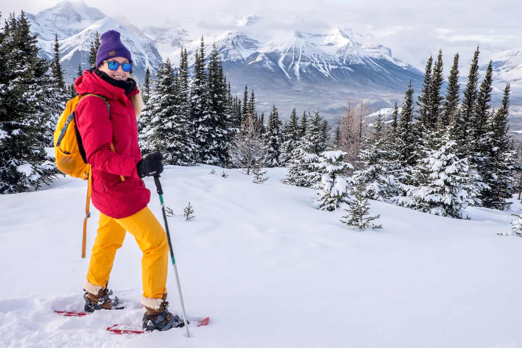 A woman in red and yellow winter clothing snowshoeing on an elevated snow-covered, forested hill overlooking a long ridge of snow-capped Rocky Mountains. 