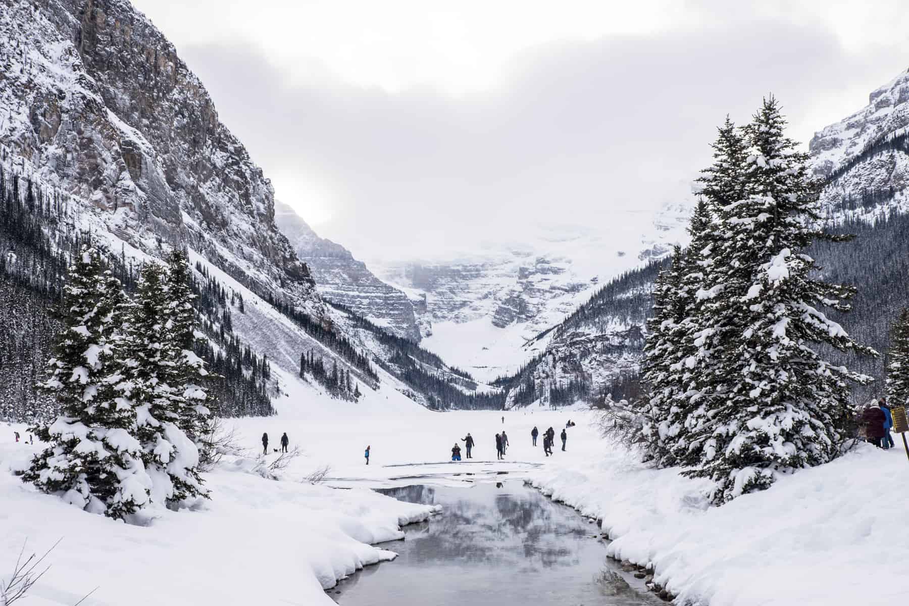 People standing on the frozen Lake Louise in Banff, surrounded by mountains, with a melted section of water in foreground. 