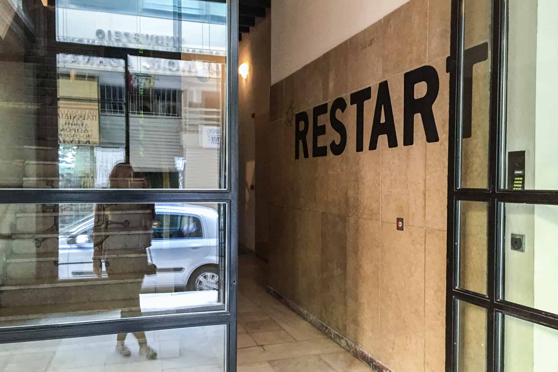 A woman's reflection in seen in the glass window of a building whose entrance hall wall has the word RESTART painted on it. 