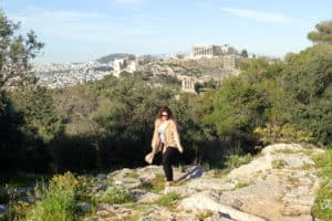 A woman walks across rocks on a green hilltop with the Acropolis of Athens in the background.