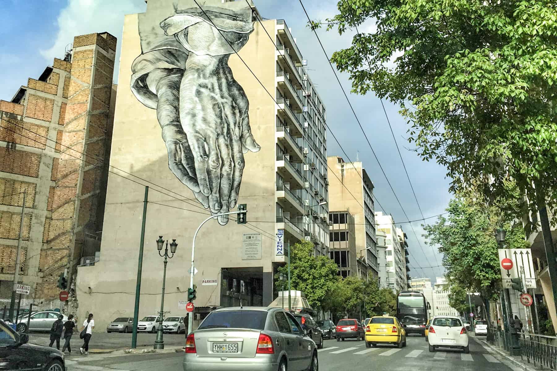 Cars driving past street art in Athens of giant praying hands on a residential building.