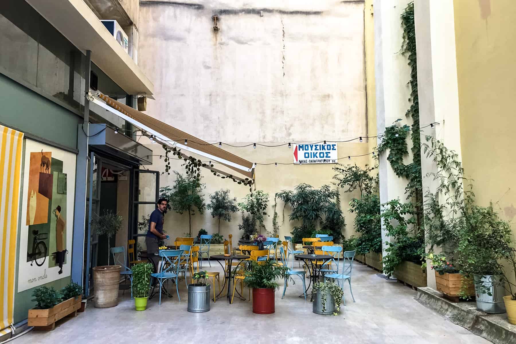 A man stands outside a cafe with blue and yellow chairs, in a quiet alleyway in Athens filled with plants. 