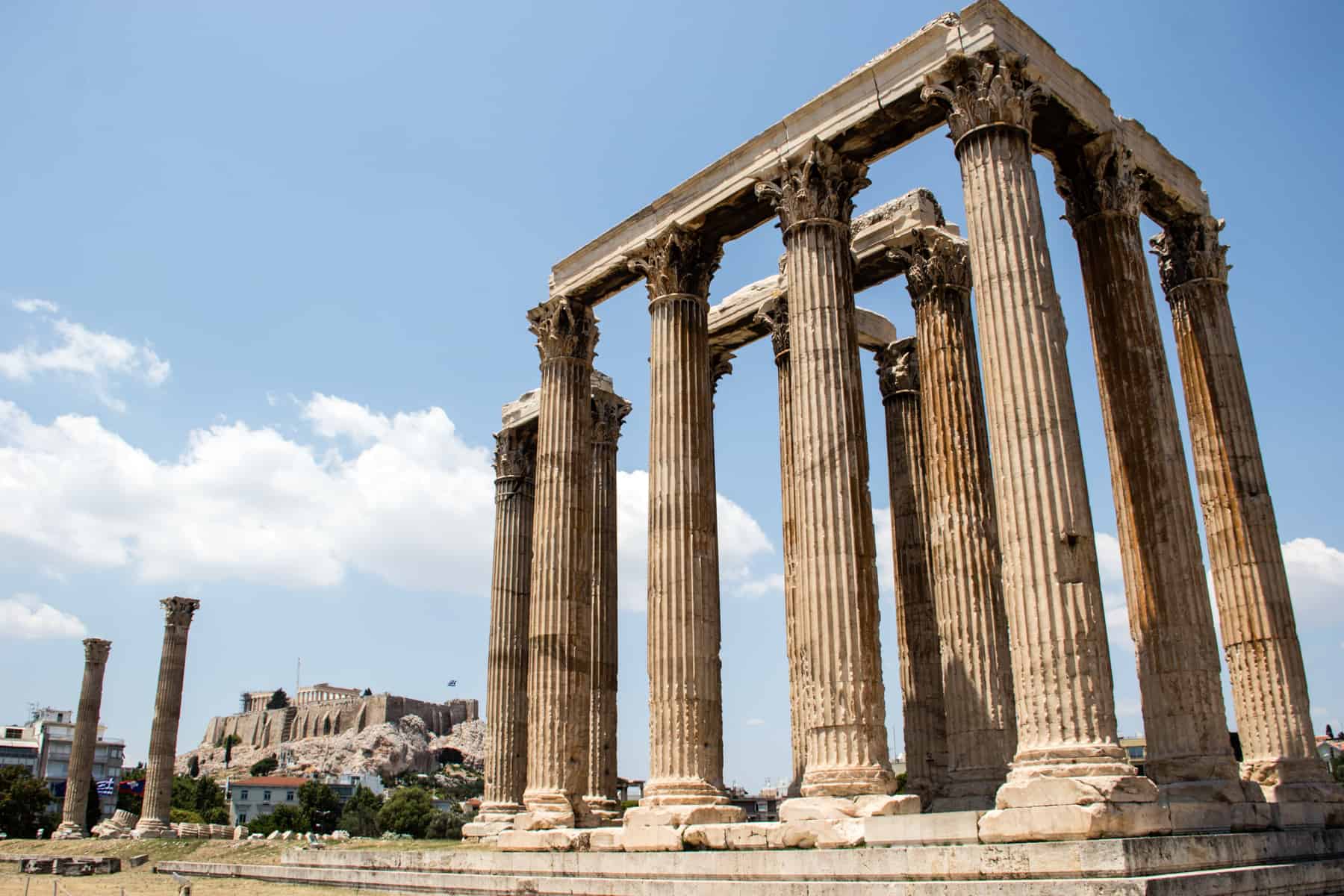 A tall, golden columned rectangular temple known as The Temple of Olympian Zeus - one of the ancient sites in Athens. The Acropolis can be seen in the background. 