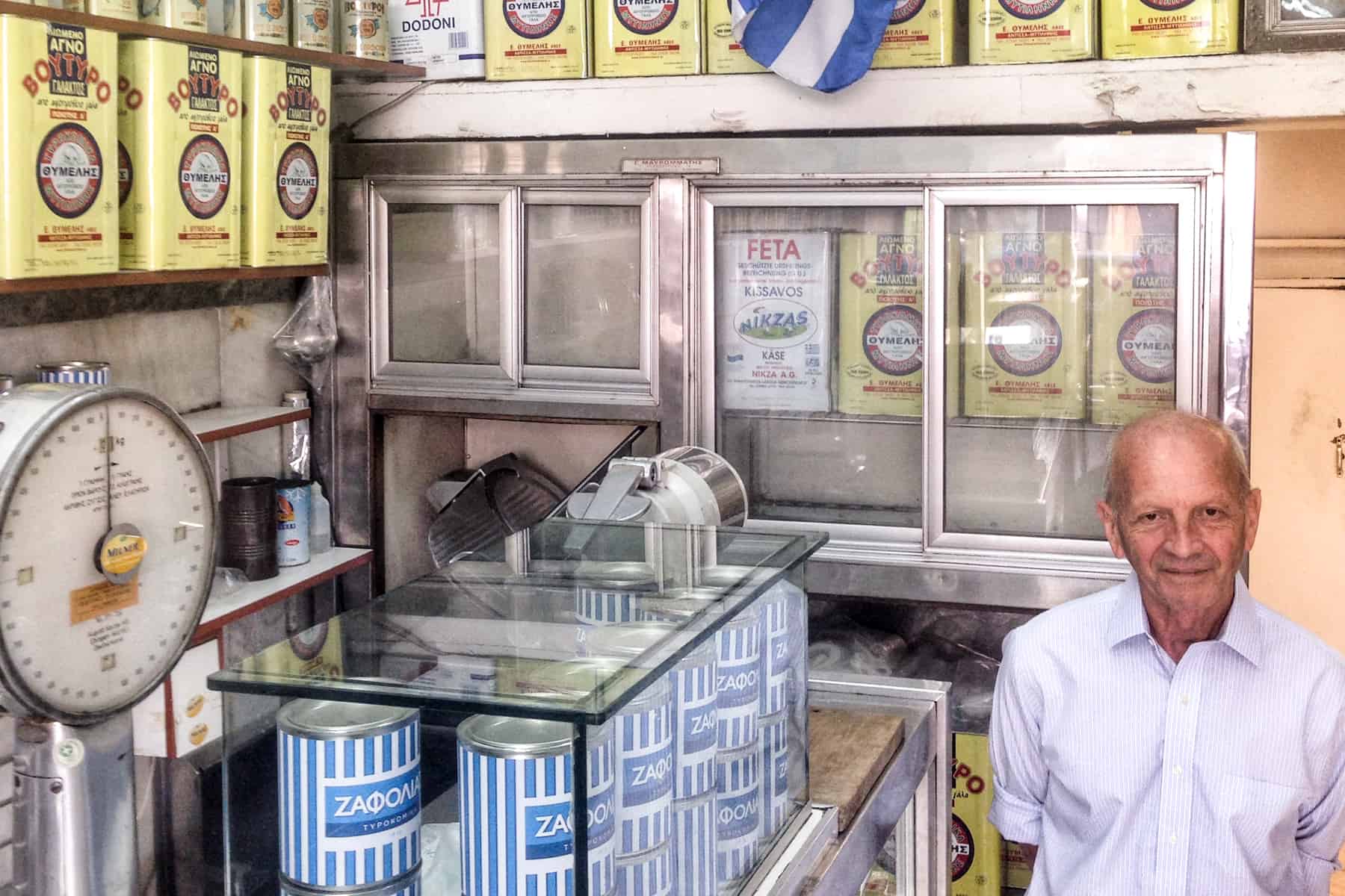 A man stands in an old store full of yellow and blue and white stripped cans of feta cheese and oil. Next to the glass strand is an old weighing scale. 