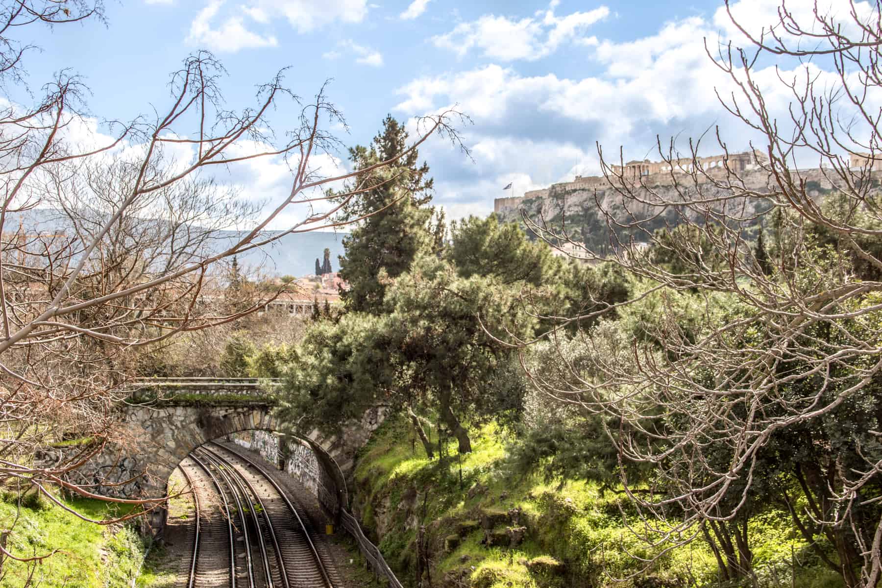 A train track cuts through a forest on the left next to the Parthenon temple on the Athenian Acropolis hill on the right.