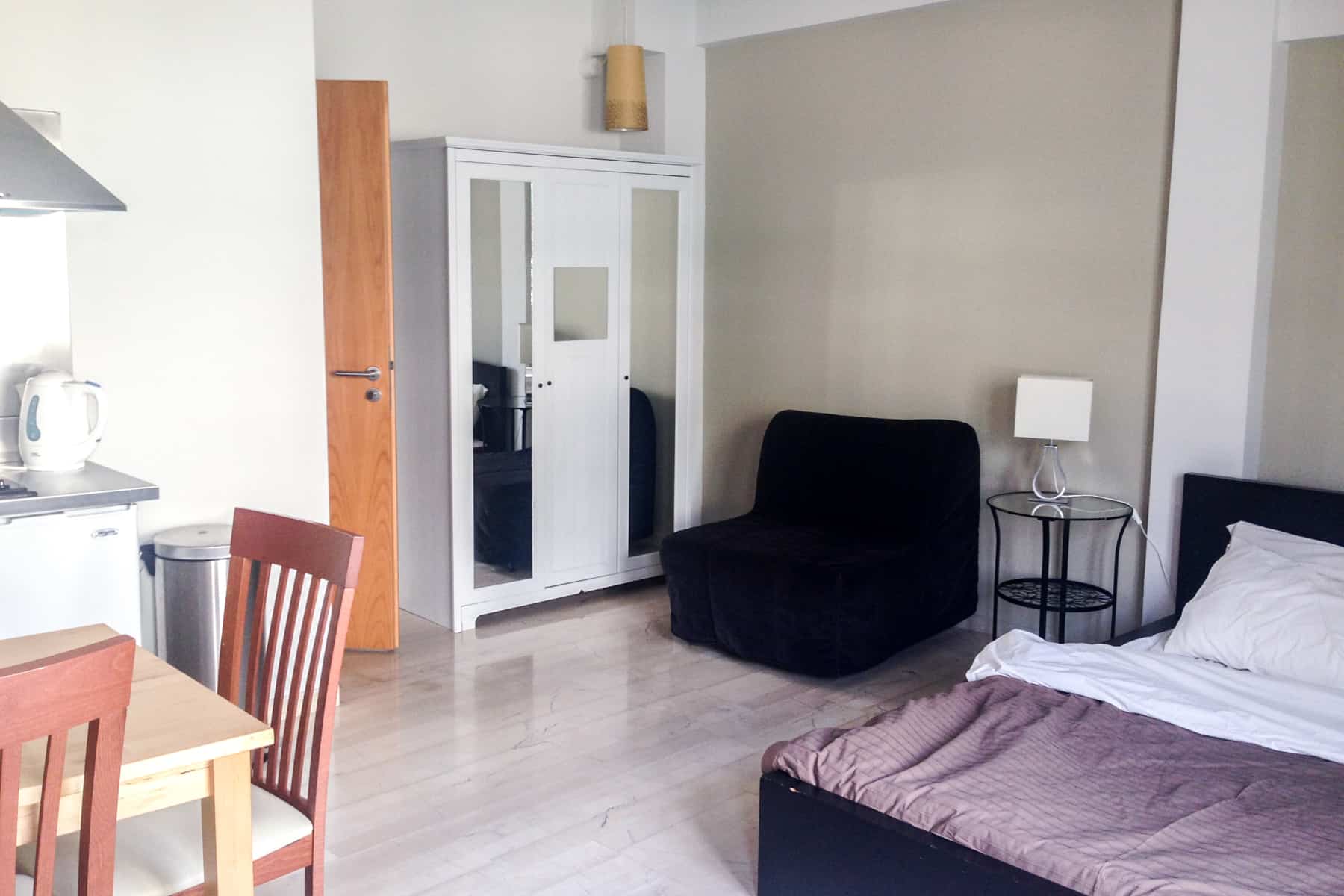 A studio apartment in Athens with (left to right) a small white kitchen and light wooden table, a white wardrobe and black single seat soft and a black bed with white and purple sheets. 
