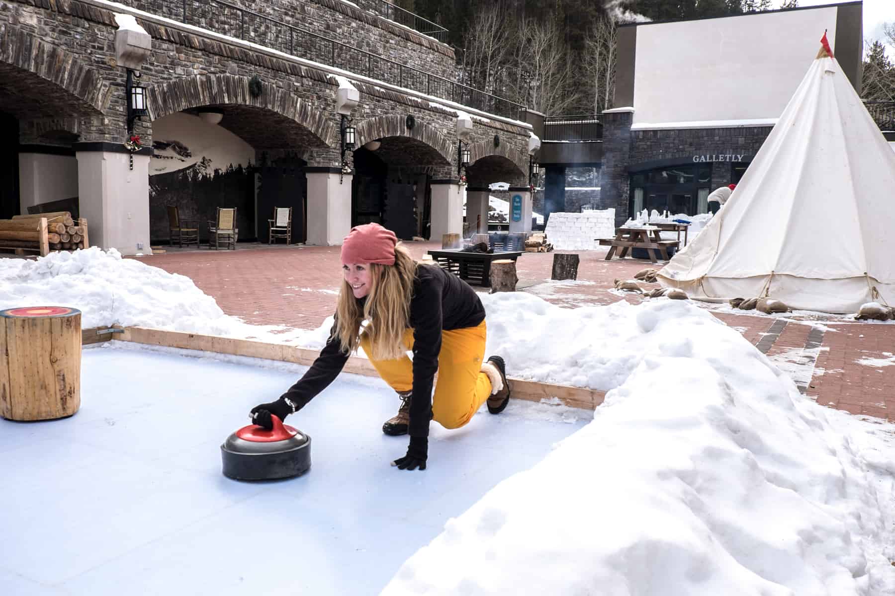 A woman dressed in black and yellow, and wearing a pink hat tries curling on a small ice rink in Banff, Canada. 