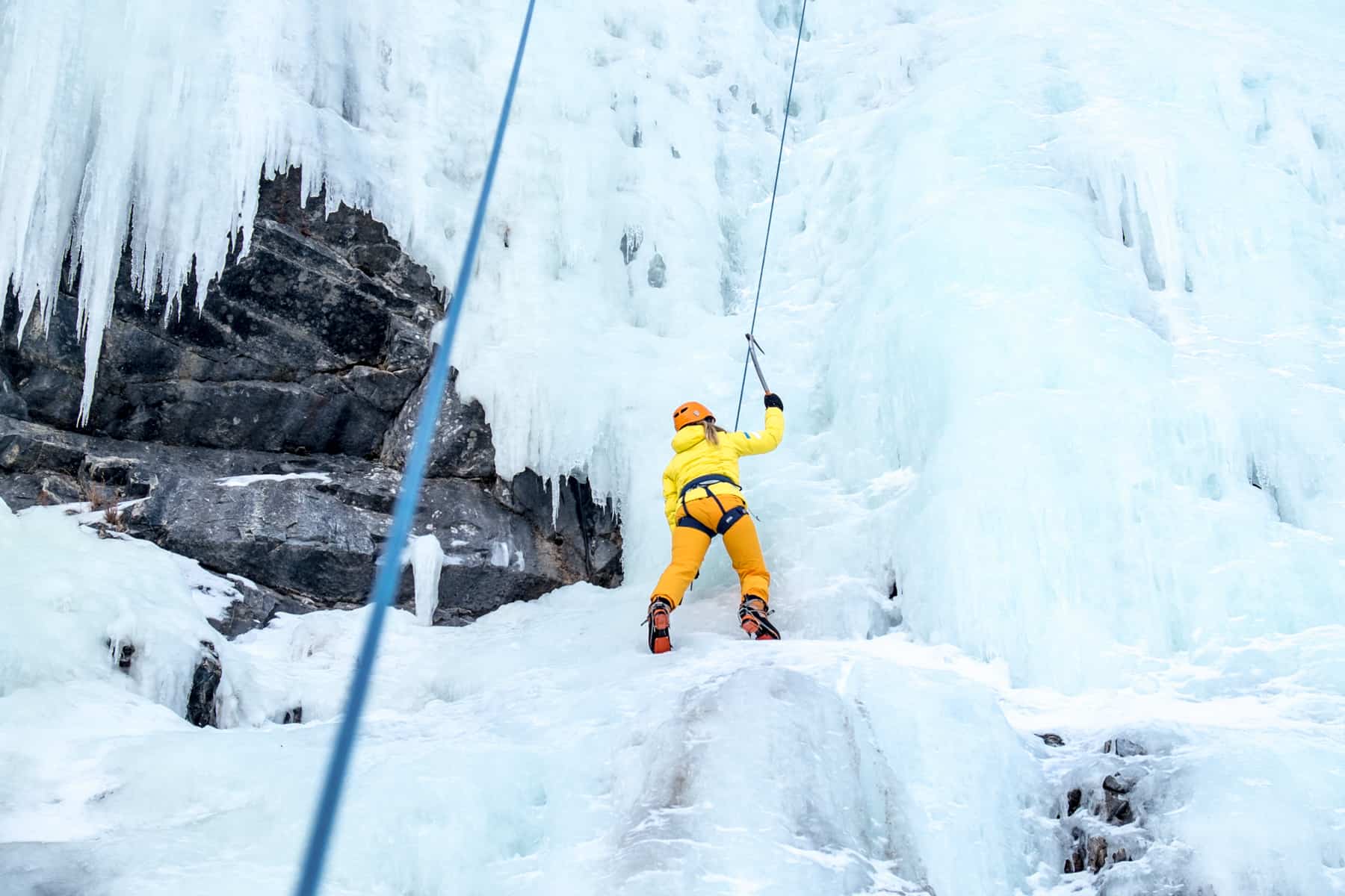 A woman in yellow and orange clothing ice climbing a frozen waterfall in Banff National Park in winter.