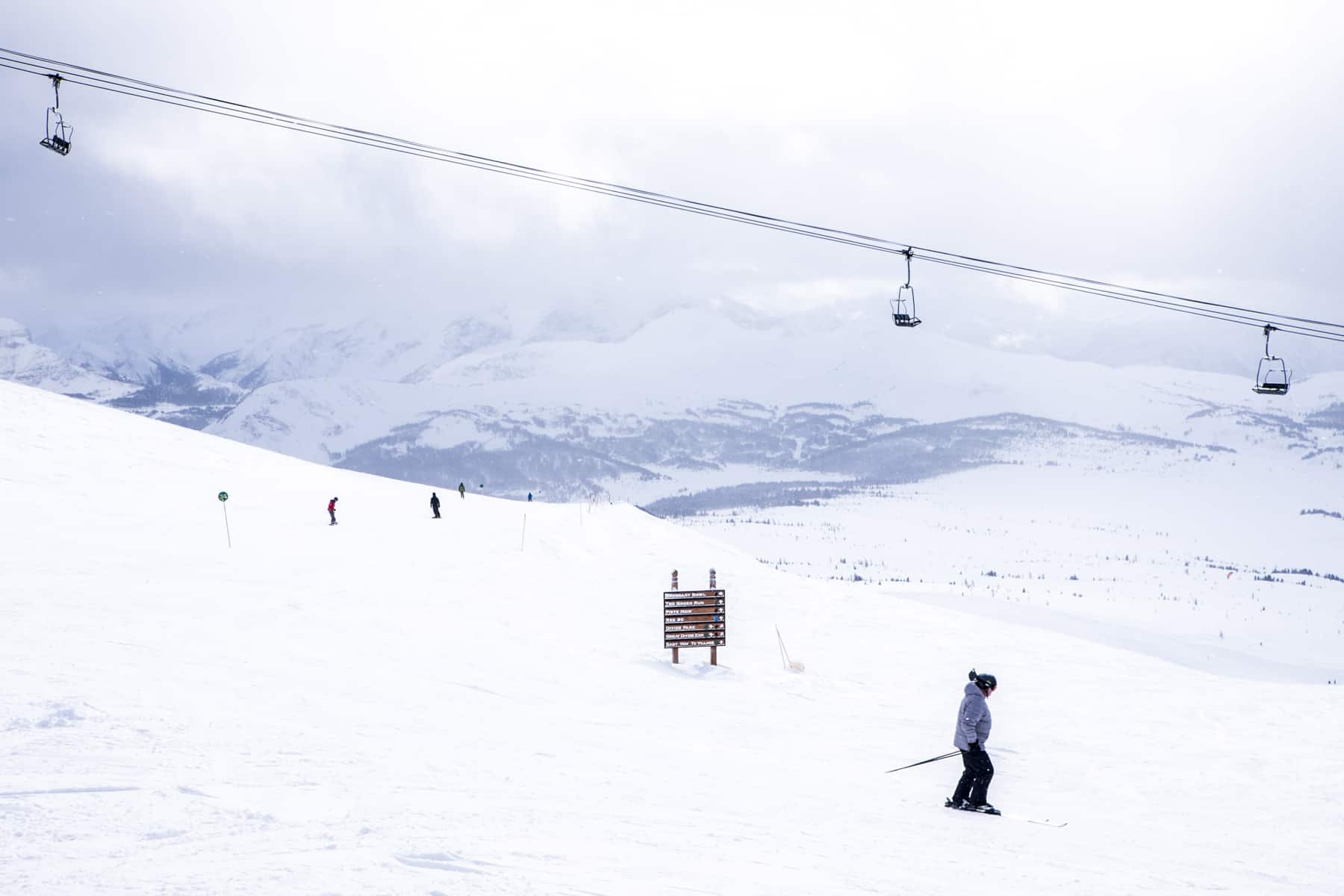 A woman skis past a wooden sign on a wide, thick snow covered slope to a backdrop of snowcapped Rocky Mountains in Banff. 
