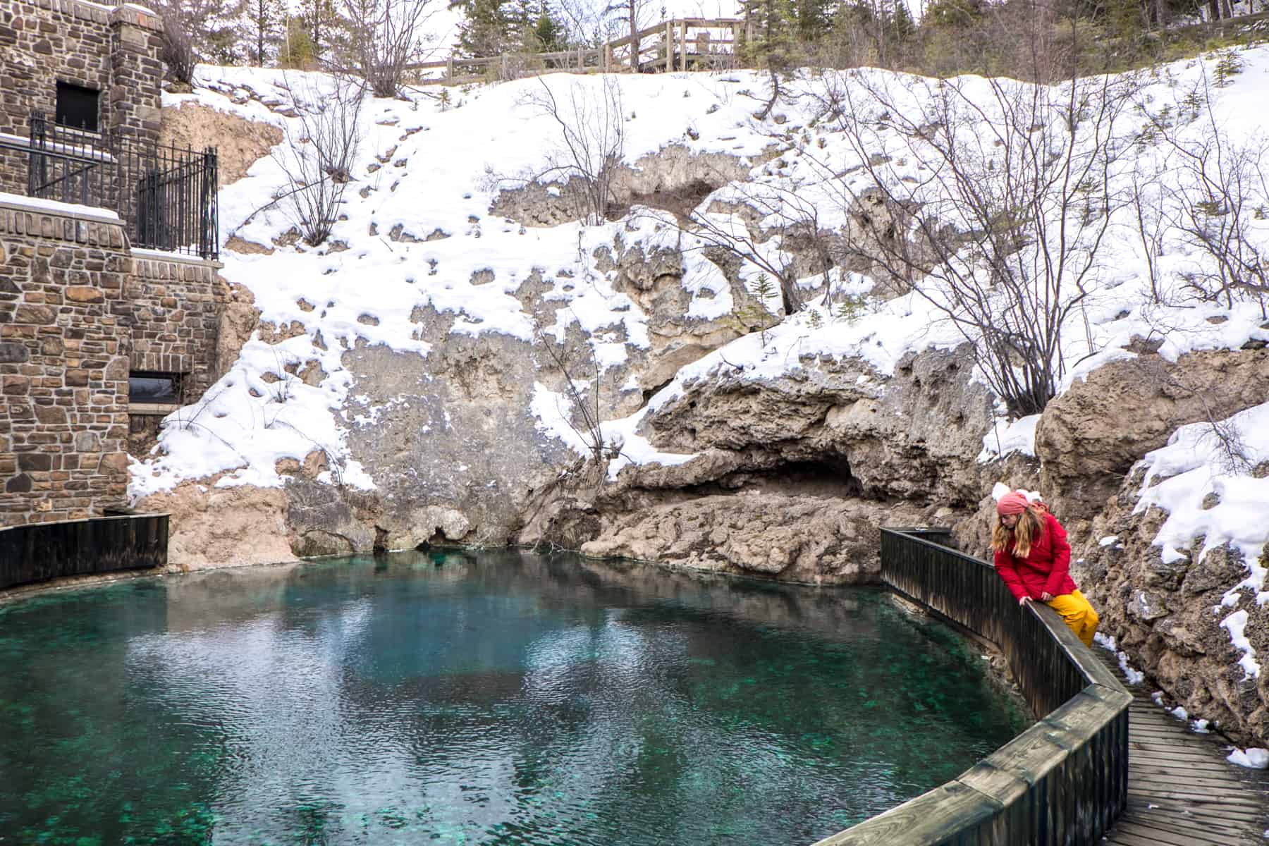 A women dressed in red and yellow looks down into a basin of water next to a snow covered wall - the hot springs of Banff in winter. 