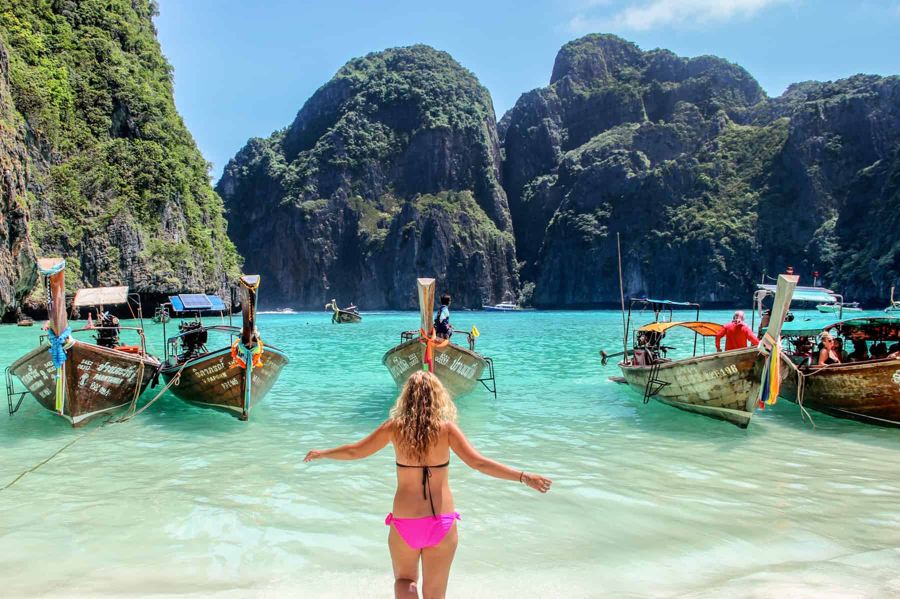 A woman in a black and pink bikini stands in the shallow clear waters of Maya Bay facing a row of five wooden boats and the surrounding limestone rocks.