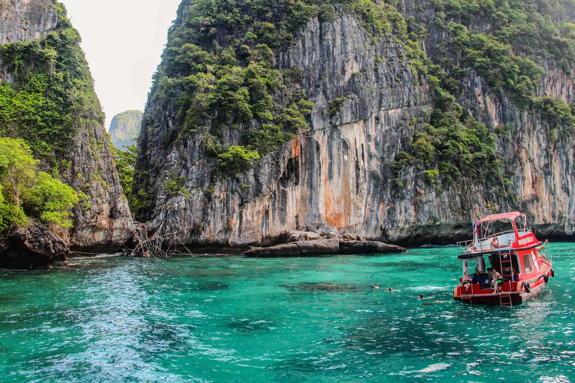 A red boat floats at an angle on bright green-blue water while people swim towards the opening between the two grass covered cliff faces.