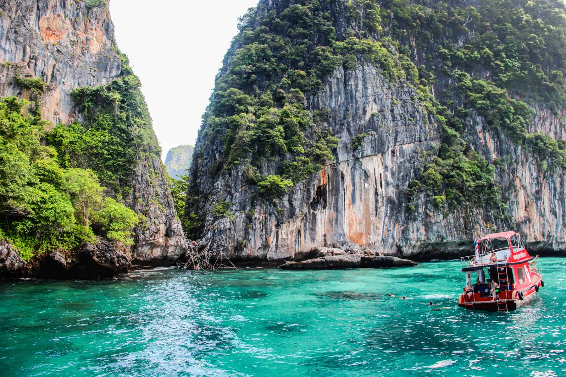 A red boat floats at an angle on bright green-blue water while people swim towards the opening between the two grass covered cliff faces.