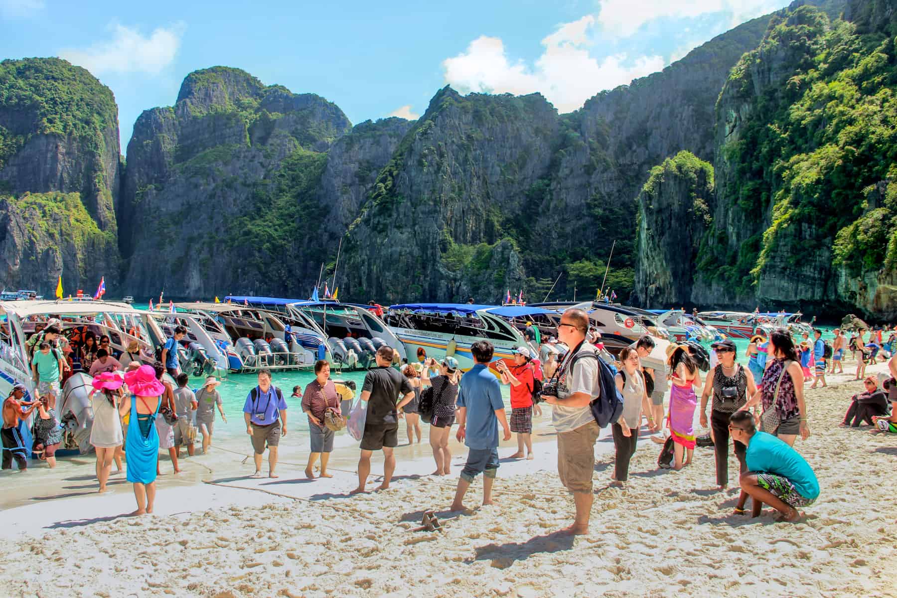 A large crowds of tourists on the white sand beaches of May Bay in Thailand, entering and exiting boats on the shoreline. The beach is surrounded by high rocks. 