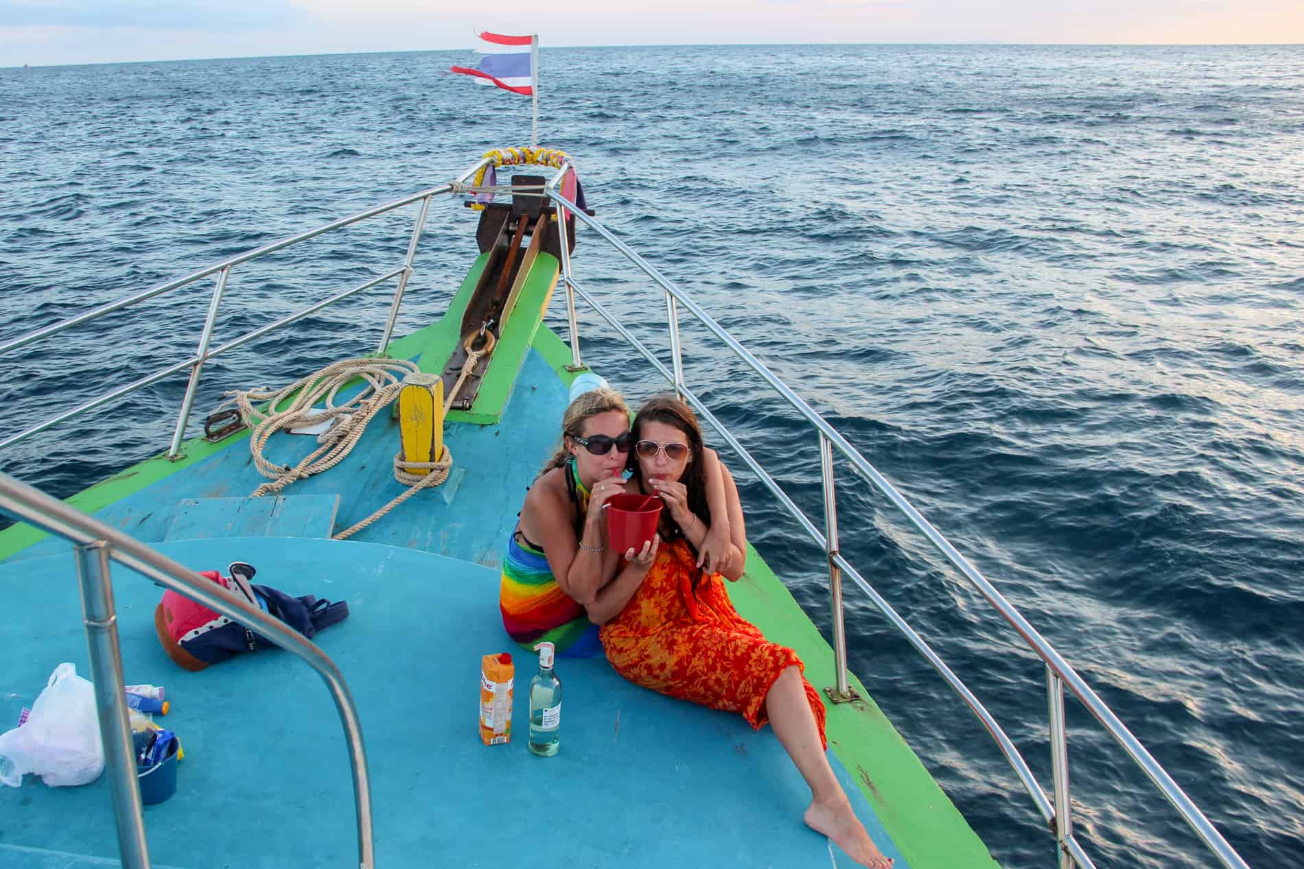 Two women in sarongs drinking through straws from a red bucket while on deck of a boat in Thailand. 