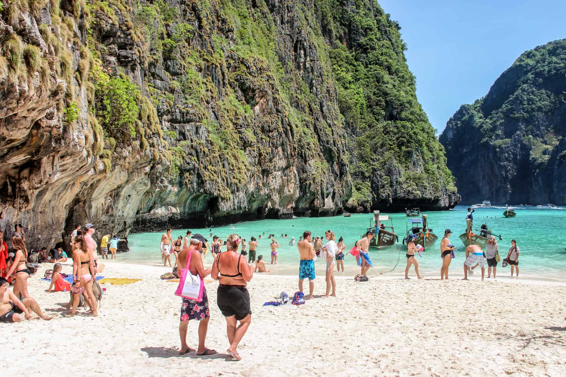A small crowd on people on the beach and swimming in the waters of the rocky cove of Maya Bay in Thailand. 